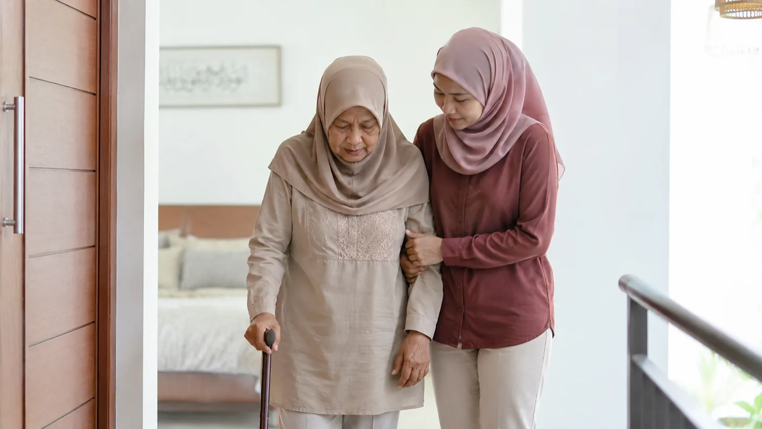 A daughter gently supporting her elderly mother as she walks with a cane at home