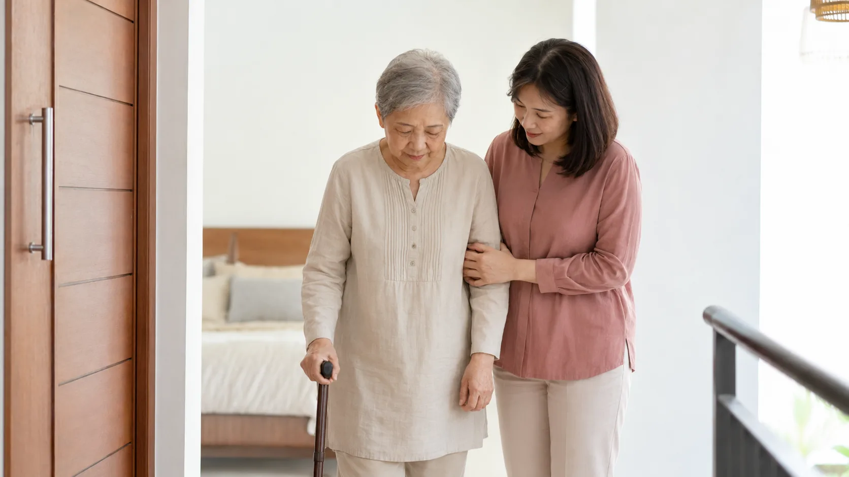 A daughter gently supporting her elderly mother as she walks with a cane at home